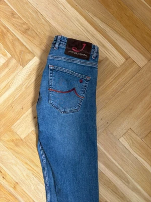 Blå jeans från Jacob Cohen - Snygga blå jeans från Jacob Cohen med klassisk femficksmodell och raka ben. Jeansen har en unik detalj med röd söm på bakfickan och patch med logga bak i midjan. Tillverkade i mjukt denimtyg som ger en skön passform.