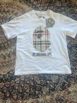  BAPE t-shirt - Säljer en vit t-shirt från A Bathing Ape med det klassiska apansikte-trycket i rutigt mönster på bröstet och texten 'BY BATHING APE' under. T-shirten har korta ärmar och rund hals. Perfekt för dig som gillar streetwear och vill sticka ut. Priset kan diskuteras vid snabb affär.