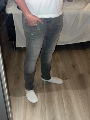Grå slim fit jeans med slitningar - Säljer ett par grå slim fit jeans med lätt slitna detaljer och diskreta färgskiftningar. Jeansen har klassisk femficksmodell och smal passform som sitter tajt längs benen. Perfekta för en avslappnad och trendig look. Finns en liten defekt i midjan, men de är inget som påverkar passformen tycker jag inte. Och de är i ett bra skicka. Har av er vid funderingar