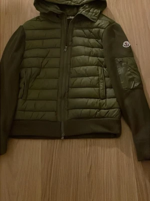 Grön cardigan   från Moncler - Stilren grön pufferjacka från Moncler med huva och quiltad front. Jackan har ribbade muddar, dragkedja framtill och en ficka med dragkedja på ärmen. Moncler-logga på vänster ärm. Perfekt för kalla dagar och riktigt snygg till streetwear.