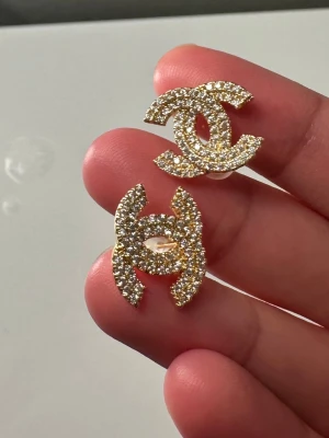 Guldiga örhängen med strass - Säljer ett par glittriga örhängen i guldton med massor av små, gnistrande stenar. Örhängena har en ikonisk dubbel-C-form och ger en lyxig känsla till din look. Perfekta för dig som vill sticka ut och addera lite extra bling till outfiten.