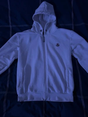 Vit hoodie med dragkedja från Moncler - Snygg vit hoodie från Moncler med dragkedja framtill och två fickor. Klassisk huva med snörning och Moncler-logga på bröstet. Tillverkad i mjukt bomullsmaterial som känns skönt mot huden. Perfekt för en clean och stilren look. 