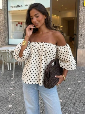 Offshoulder prickig topp med puffärm - Superfin offshoulder topp i vitt med svarta prickar och puffiga trekvartsärmar. Toppens vida passform och luftiga material gör den perfekt för varmare dagar. Snygg att styla med jeans eller kjol för en trendig look.