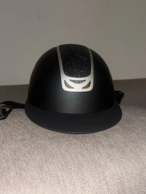 Svart ridhjälm med glitterdetalj - Snygg svart ridhjälm från Horze med en glittrig detalj och silverfärgad kant på ovansidan. Den är dock något repig på vissa ställen men funkar helt som den ska och inget som syns jätte mycket.  Hjälmen har justerbart hakband och mjuk vaddering på insidan för extra komfort. Perfekt för dig som vill ha både stil och säkerhet i stallet.