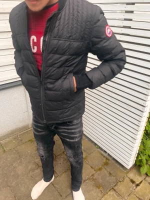 Svart pufferjacka från Canada Goose - Säljer en svart pufferjacka från Canada Goose med quiltad design och diskret logga på ärmen. Jackan har dragkedja framtill, ståkrage och två sidofickor. Perfekt för kalla dagar och riktigt snygg till streetwear-stilen.