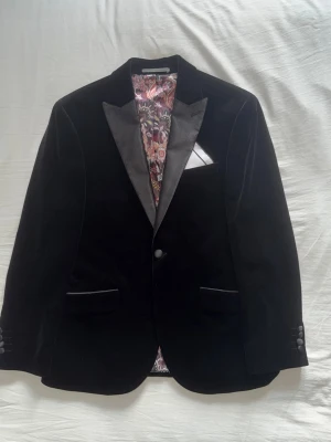 Next Signature italian fabric slim fit velvet blazer - kavaj 48S - Stilren svart kavaj i sammet med breda slag och två knappar framtill. Insidan har ett färgstarkt blommigt foder i rosa, lila och beige toner. Klassisk ficknäsduk i bröstfickan och flera innerfickor. Perfekt för dig som vill sticka ut lite extra. Next Signature i stl 48S slim fit. Original pris: 2920kr  Skriv för mer detaljerad information eller bilder :)
