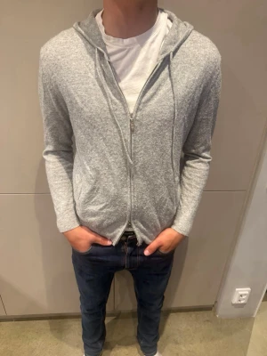 Ljusgrå zip kofta av kashmir  - En enkel och stilren ljusgrå ziptröja med huva och dragskedja. Tröjan har långa ärmar, två fickor framtill och är gjord i ett mjukt, tunt kashmir som är varm och skön men samtidigt bra för sommarkvällar och hösten. Perfekt att bära över en t-shirt för en avslappnad look.
