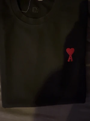 Svart t-shirt från Ami Paris - Svart t-shirt från Ami Paris med rund hals och en liten röd broderad logga med hjärta och bokstaven A på bröstet. Enkel och stilren design som passar till det mesta. Tillverkad i mjuk bomull för skön känsla.