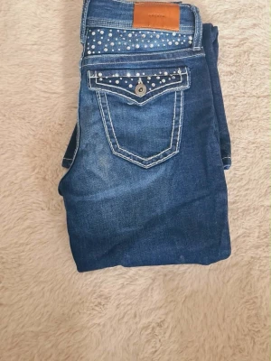  bootcut jeans med rhinestone detaljer - Snygga blå bootcut jeans med rhinestones på bakfickan och kontrastsömmar. Jeansen har en dekorativ ficka med knapp och läderpatch i midjan. Perfekta för dig som gillar en retroinspirerad look och vill sticka ut lite extra💕 strl 152
