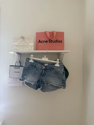 Blå denimshorts med broderi - Säljer ett par ljusblå denimshorts med råa kanter och snygga slitningar framtill. På bakfickorna finns broderade fjärilar som ger en unik touch. Shortsen har klassisk femficksdesign och knappgylf. Perfekta för sommardagar! Har xs och passar bra på mig