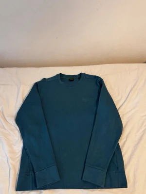 Blå sweatshirt från BOSS - Snygg blå sweatshirt från BOSS med diskret logga broderad på bröstet. Tröjan har rund halsringning, långa ärmar och breda muddar vid ärmslut och nederkant. Använd 2 gånger endast