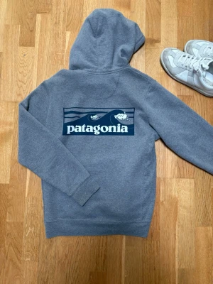 Grå Patagonia hoodie med tryck - Snygg grå hoodie från Patagonia med stor logga och bergsmotiv på ryggen samt liten logga på bröstet. Klassisk känguruficka framtill och justerbar huva med dragsko. Mjuk och skön bomullsblandning, perfekt för chill dagar.  Nypris 1100kr, mitt pris 499kr! Pris går att diskutera! S i man, M i kvinna