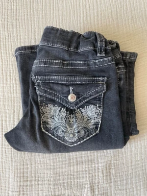 Mörkblå bootcut jeans med brodyr - Snygga mörkblå jeans med bootcut-modell och dekorativ brodyr på bakfickan. Jeansen har vita kontrastsömmar och en knappstängning fram. Materialet ser ut att vara klassisk denim med lite stretch för extra komfort.