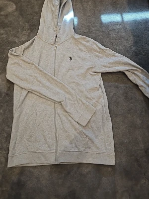 Ljusgrå ziphoodie från U.S. Polo Assn. - Ljusgrå hoodie med dragkedja och huva. Har en liten mörk brodyr på bröstet med en polospelare. Tillverkad i mjukt bomullsmaterial och har fickor framtill. Perfekt för en chill och avslappnad stil. Passar perfekt för dig som har storlek 170 till 176.