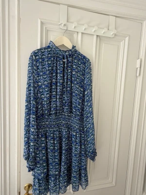 Blåvit mönstrad kortklänning - Kortklänning med blått och vitt grafiskt mönster, hög krage och långa ärmar. Klänningen har smock i midjan och ett luftigt, flowy tyg som ger en avslappnad siluett. Perfekt för dig som gillar färg och mönster.