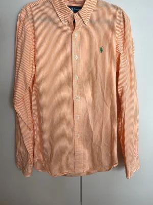 Randig skjorta från Ralph Lauren - Säljer en orange och vit randig skjorta från Ralph Lauren i slim fit. Skjortan har klassisk krage, långa ärmar och knappar framtill. Broderad grön logga på bröstet. Tillverkad i mjuk bomull, perfekt för en clean och stilren look.
