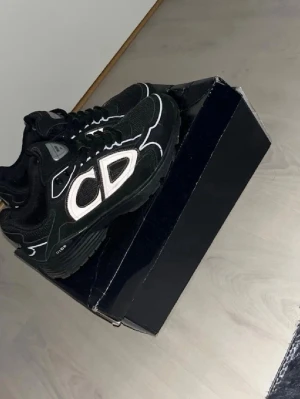 Svarta sneakers med CD-detalj - Snygga svarta sneakers med stora CD-detaljer på sidan och vita samt grå kontrastsömmar. Skorna har en chunky sula och meshpaneler för extra andningsförmåga. Perfekta för dig som gillar streetwear och vill sticka ut med en unik design.