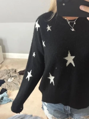 Svart stickad tröja med stjärnor - Svart stickad tröja med vita stjärnor i olika storlekar över hela tröjan. Tröjan har en avslappnad passform och långa ärmar. Perfekt för dig som gillar en cool och trendig look.