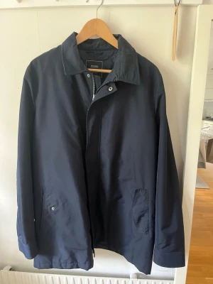 Mörkblå klassisk rock med krage - Stilren mörkblå rock med enkel knäppning och klassisk krage. Två snedställda fickor framtill och diskret detalj på ärmen. Passar perfekt till både jeans och kostym. Materialet känns slätt och lätt, vilket gör den enkel att bära under olika säsonger. Rocken/jackan är från Boomerang 
