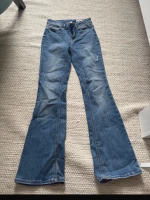 Blå bootcut jeans från Vero Moda - Säljer ett par klassiska blå bootcut jeans från Vero Moda. Jeansen har normal midja, fem fickor och lätt utsvängda ben. Tillverkade i mjuk denim med snygga slitningar framtill. Perfekta för en avslappnad och trendig look.