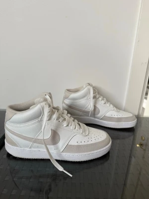 Vita Nike sneakers  - Säljer ett par vita nike sneakers med beige detaljer och klassisk snörning. Skorna har perforerade partier framtill för extra ventilation och en stilren design med tydlig sula. Perfekta för dig som gillar en clean och sportig look.