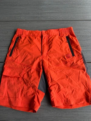 Orange funktionsshorts - Snygga orange funktionsshorts med gråa detaljer och dragkedjefickor framtill. Perfekta för aktiva dagar utomhus.