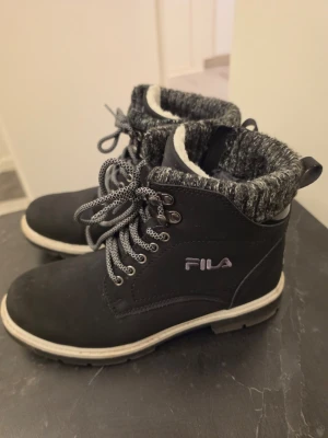 Fils Svarta kängor med foder - Snygga svarta kängor från FILA med grov sula och kontrasterande vita detaljer. Kängorna har gråmelerat, stickat foder runt skaftet och matchande snören. Perfekta för kalla dagar och ger en cool streetstil.