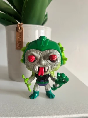 Grön och grå samlarfigur Funko pop - Cool samlarfigur med grått ansikte, röda ögon och grön hjälm. Figuren har en utstickande röd tunga, gröna detaljer på huvudet och håller i ett grönt tillbehör. Kroppen är svart, lila och turkos med vita ben. Perfekt för dig som gillar unika figurer. Funko pop