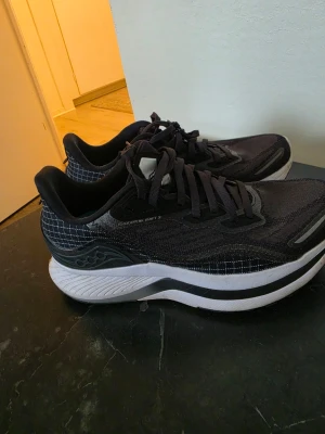 Svarta sneakers  - Snygga svarta sneakers från Saucony med vita och grå detaljer. Ovandelen är i mesh med rutmönster och skorna har en tjock, vit sula med svarta inslag. Perfekta för dig som gillar sportig stil och vill ha bekväma skor till vardags.