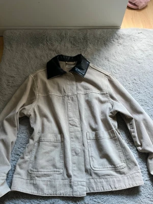 Beige overshirt från Perfect Jeans - Stilren beige overshirt från Perfect Jeans med två stora framfickor och en svart krage i skinnimitation. Overshirten har raka linjer och en avslappnad passform, perfekt att slänga över en t-shirt för en cool look.