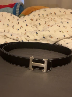 Svart skärp med silverfärgat H-spänne - Snyggt svart bälte med ett stort, silverfärgat H-format spänne. Bältet har vita sömmar längs kanterna som ger en stilren kontrast. Perfekt accessoar för att lyfta din outfit.
