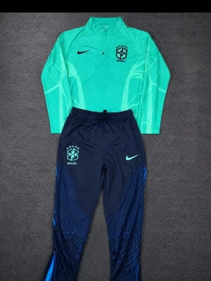 Brasilien Nike träningsset - Snyggt träningsset från Nike med Brasilien-tryck. Överdelen är turkos med lång ärm och halv dragkedja, byxorna är mörkblå med turkosa detaljer och Brasilien-logga. Perfekt för fotbollsträning eller sportiga dagar.