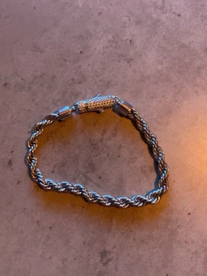 Silverfärgat tvinnat armband - Snyggt armband i silverfärgad metall med tvinnad kedja och ett lås dekorerat med små gnistrande stenar. Armbandet har en stilren och modern look som passar perfekt till olika outfits.