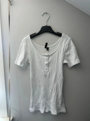 Vit ribbad topp från Pieces - Säljer en vit ribbad topp från Pieces med korta ärmar och rundad halsringning. Toppen har knappar framtill som en snygg detalj och är tillverkad i mjukt material som sitter skönt på kroppen. Perfekt till jeans eller kjol!