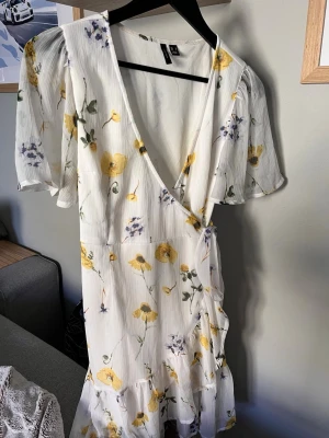 Vit blommig omlottklänning - Söt vit omlottklänning med blommigt mönster i gult och lila. Klänningen har korta, luftiga ärmar, volangkant nertill och knytband i midjan. Perfekt för varma dagar och ger en romantisk look.