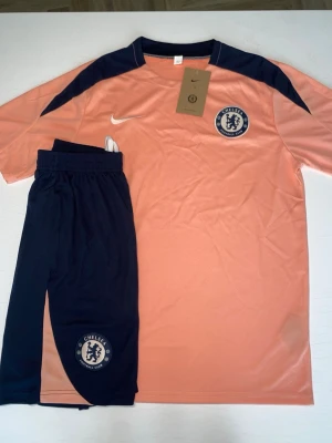 Chelsea träningsset i persika och marinblå - Säljer ett Chelsea FC träningsset från Nike i persikofärg med marinblå detaljer. Setet består av en kortärmad tröja med klubbmärke och matchande shorts med elastisk midja. Materialet är lätt och andas, perfekt för träning eller match.