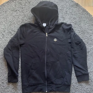 Svart zip hoodie från Moncler - Snygg svart hoodie från Moncler med dragkedja framtill och klassisk Moncler-logga på bröstet. Hoodien har huva, två stora fickor fram och ribbade muddar vid ärmslut och nederkant. Perfekt för en chill och stilren look. Priset kan diskuteras 