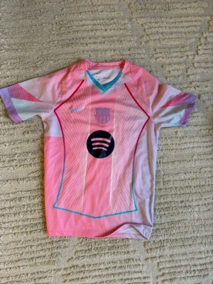 Rosa FC Barcelona - Säljer en rosa FC Barcelona matchtröja från Nike med Dri-FIT-material. Tröjan har korta ärmar, V-ringning med turkos kant och tryckta loggor från både Nike och Spotify. Perfekt för dig som älskar fotboll och vill sticka ut på planen.