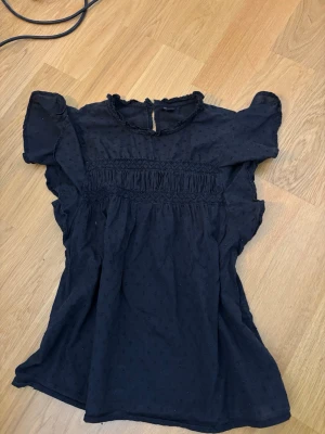 Svart blus med volangärmar - Snygg svart blus med korta volangärmar och diskret prickigt mönster. Blusen har rund halsringning med liten volangkant och knapp i nacken. Smockdetaljer framtill ger en fin passform. Perfekt till jeans eller kjol.