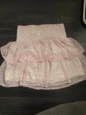 Blommig volangkjol från Vero Moda - Supersöt kort kjol från Vero Moda med volanger och blommigt mönster i rosa och vitt. Kjolen har smock i midjan för en bekväm passform och är gjord i ett lätt och luftigt material, perfekt för varma dagar.