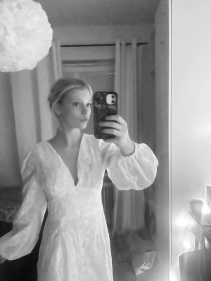 Vit långklänning med spets och v-ringning - Romantisk vit långklänning med v-ringning, puffiga långärmar och fina spetsdetaljer. Klänningen har knappar framtill och ett elegant, figurnära snitt som ger en klassisk look. Perfekt för sommarens speciella tillfällen.
