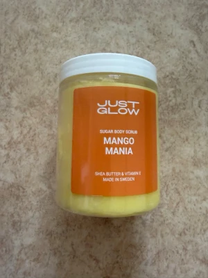 Just Glow Mango Mania Body Scrub - Sockerbaserad kroppsskrubb med doft av mango från Just Glow. Innehåller sheasmör och vitamin E för extra återfuktning. Oanvänd och plomberad. Tillverkad i Sverige. Nypris 399kr