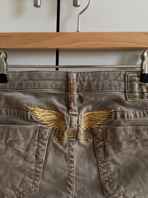 Lowrise beigegrå jeans - Äkta vintage lowrise jeans, raka och stretchiga så de sitter tajt men inte hela vägen ner. Skönt material, inte denim. Guld detaljer fram och bak och mest grå i färgen. 