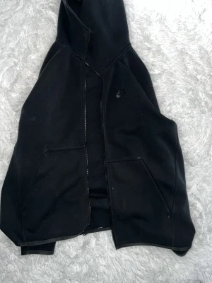 Svart Nike hoodie med dragkedja - Säljer en svart hoodie från Nike med dragkedja framtill och två stora fickor. Hoodien har en diskret Nike-logga på bröstet och är tillverkad i ett mjukt och bekvämt material. Perfekt för en avslappnad och sportig stil.