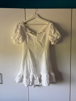 Vit broderad klänning med puffärm - Söt vit klänning med broderade blommor, puffärmar och volangkant nertill. Klänningen har en öppen rygg med knytband och små pärldetaljer längs urringningen. Perfekt för sommarens alla tillfällen!