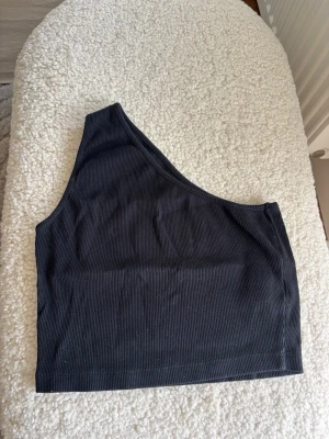 Svart oneshoulder ribbad topp - Säljer en svart ribbad topp med oneshoulder-design. Toppen är croppad och har en stilren look som passar perfekt till jeans eller kjol. Materialet är mjukt och stretchigt för en bekväm passform.