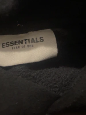 Svart Essentials Fear of God hoodie Storlek L - Säljer en svart hoodie från Essentials Fear of God. Hoodien har en klassisk huva och mjukt material på insidan för extra komfort. Diskret Essentials-logga på bröstet och ärmen ger en clean och stilren look. Perfekt för dig som gillar streetwear och vill ha något bekvämt.