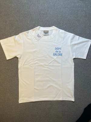 Vit t-shirt från Gallery Dept. - Snygg vit t-shirt från Gallery Dept. med blå text 'DÉPT. de la GALERIE' tryckt på bröstet. Klassisk passform och rund hals. Tillverkad i mjuk bomull som känns skön mot huden. Perfekt för en clean och stilren look.