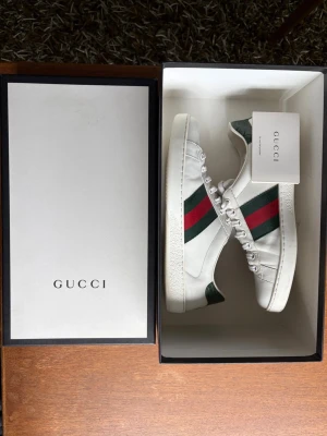 Gucci Ace Sneakers - skor + skolåda - Gucci ace sneakers i storlek 41,5. Orginial Gucci-box medföljer vid köp. Klassiska vita lädersneakers med Guccis gröna/röda Web Stripe och grön krokodilpräglad häl.