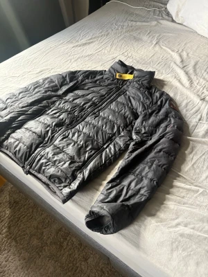 Svart dunjacka med gul detalj - Snygg svart dunjacka med quiltad design och hög krage. Jackan har dragkedja framtill, två sidofickor och en gul detalj vid halsen. Perfekt för kalla vinterdagar och riktigt lätt att bära.